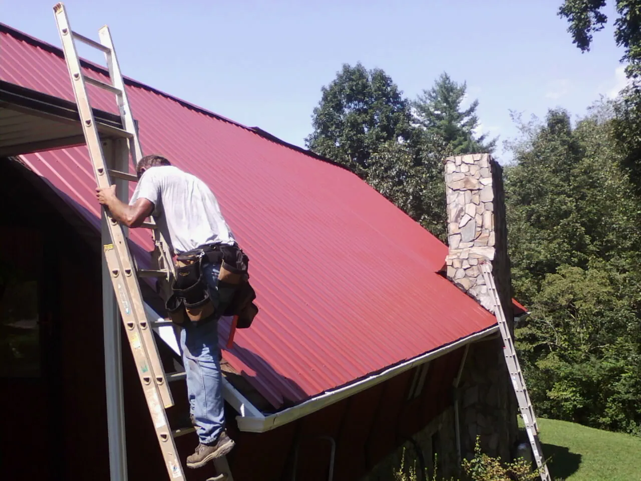 Roofer on a ladder beside a red metal roof and stone chimney — Soffit & Fascia Repair by Roofing Brothers in Lockport